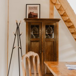 Jola Igielska - Rustykalne wnętrze: drewniana szafa z przeszkleniami, krzesło z plecionym siedziskiem, wieszak na ubrania i schody z belek, plakat 'Chatka Gorzystów' na szafie.