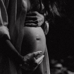 Jola Igielska - Czarno-białe zdjęcie profilowe kobiety w ciąży, z rękami obejmującymi brzuch, w rozpiętej koszuli, z delikatnym oświetleniem podkreślającym kształt brzucha.