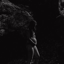 Jola Igielska - Czarno-białe zdjęcie kobiety w ciąży, stojącej boso w otoczeniu skał i liści, z rękami na brzuchu, w naturalnym oświetleniu.