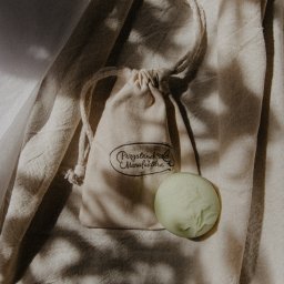 Jola Igielska - Naturalne mydło w pastelowym odcieniu zieleni leży obok lnianego woreczka z logo 'Przystanek Manufaktura' na tle beżowej tkaniny w cieniu.