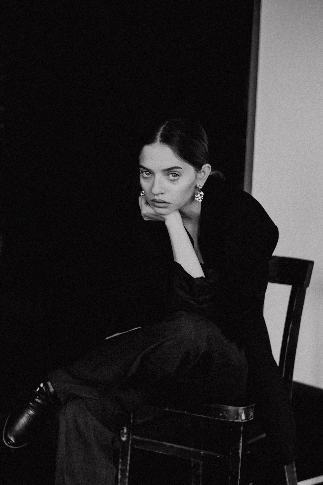 Portret kobiety siedzącej na krześle, w czarnej marynarce i spodniach, z ręką podpartą o brodę, w dużych kolczykach na tle czarno-białym.