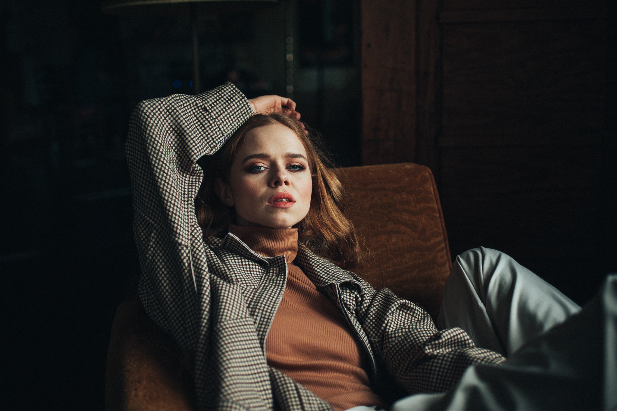 Portret kobiety w kraciastej marynarce, leżącej w fotelu z ręką za głową, z delikatnym makijażem i rozpuszczonymi włosami, w ciemnym wnętrzu.