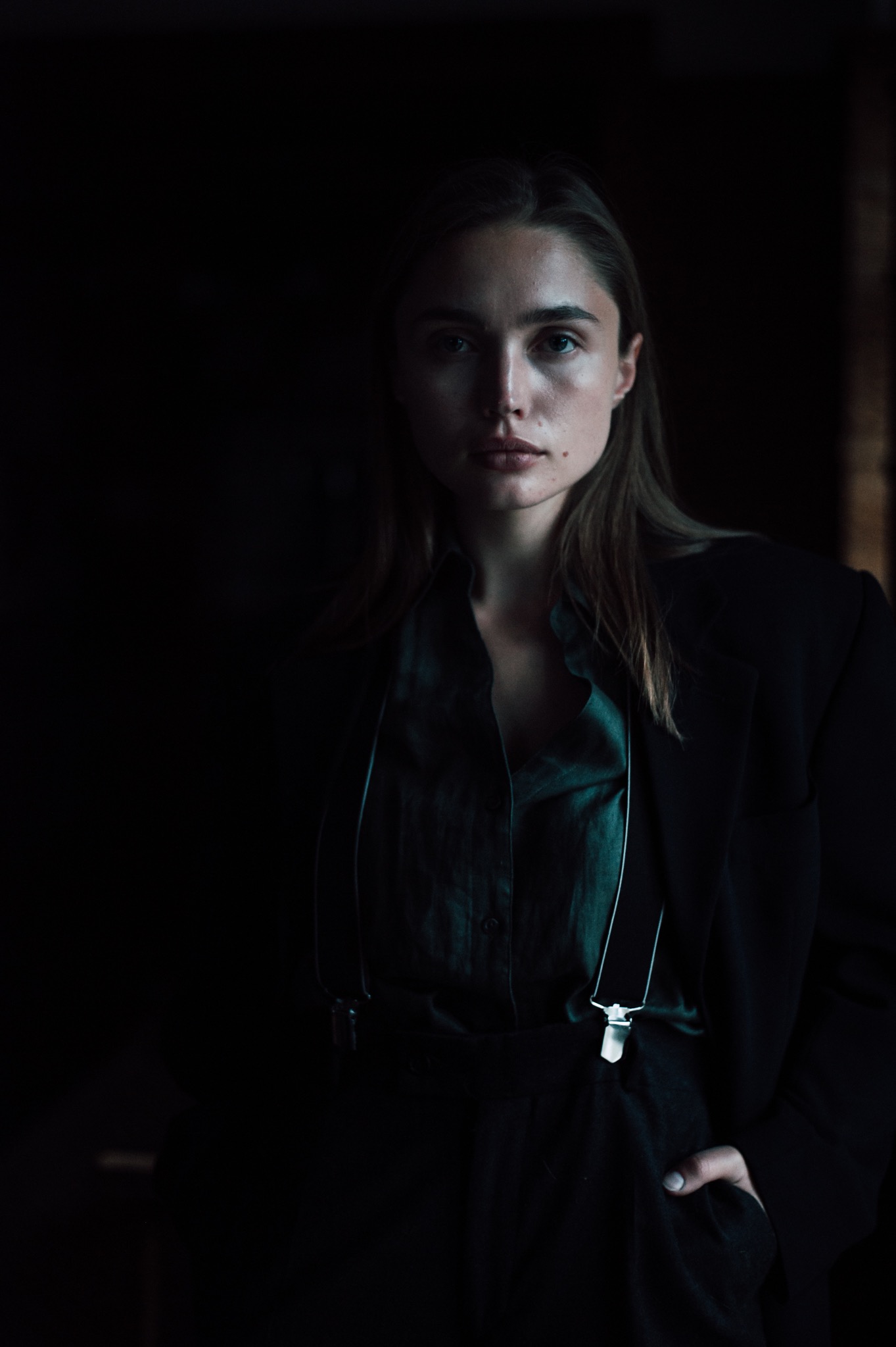 Portret kobiety w ciemnym garniturze z szelkami, oświetlonej z przodu, z ręką w kieszeni.