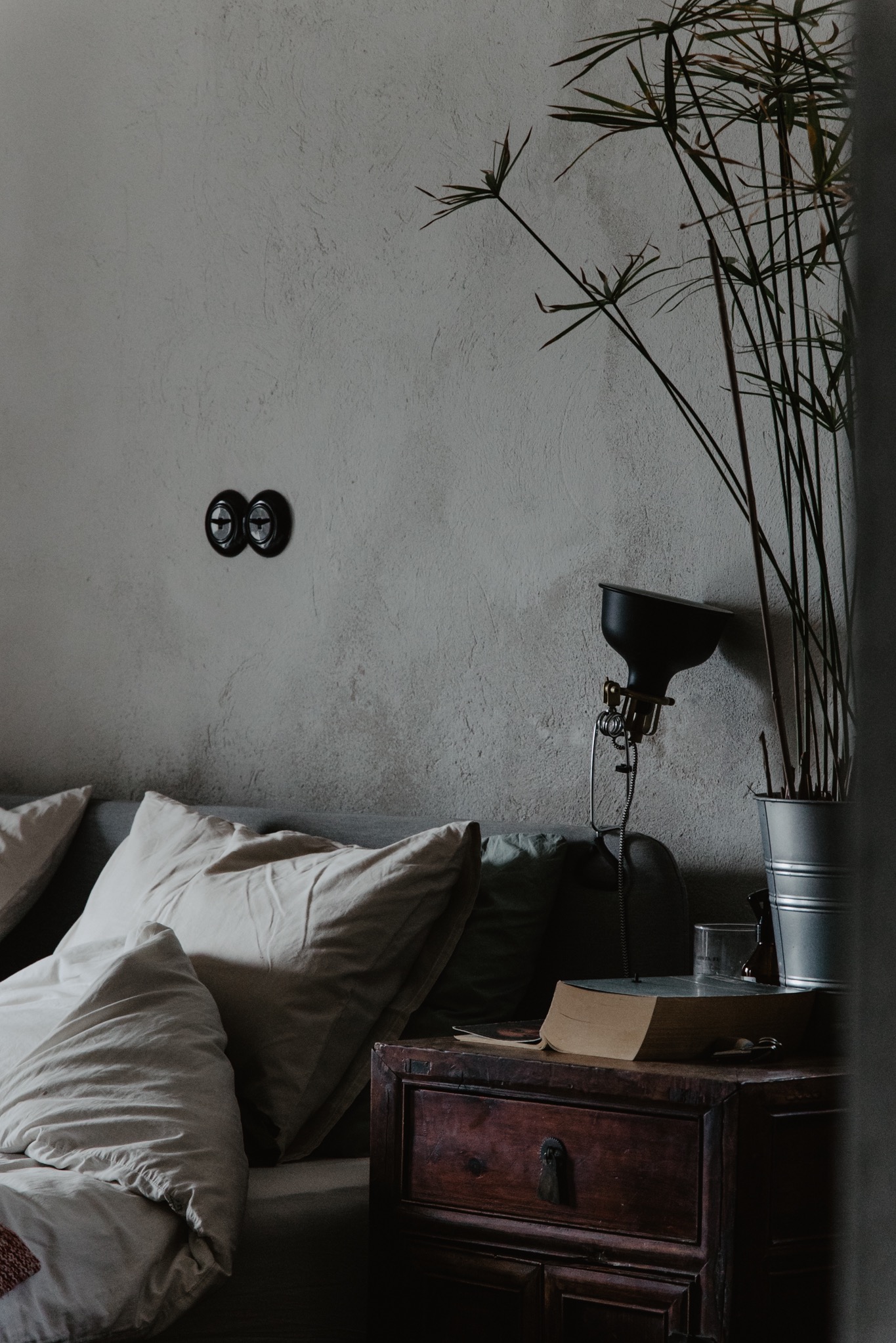 Minimalistyczna sypialnia: łóżko z jasną pościelą, drewniana szafka nocna z książką i lampką, roślina w metalowej doniczce na tle ściany z włącznikami światła.