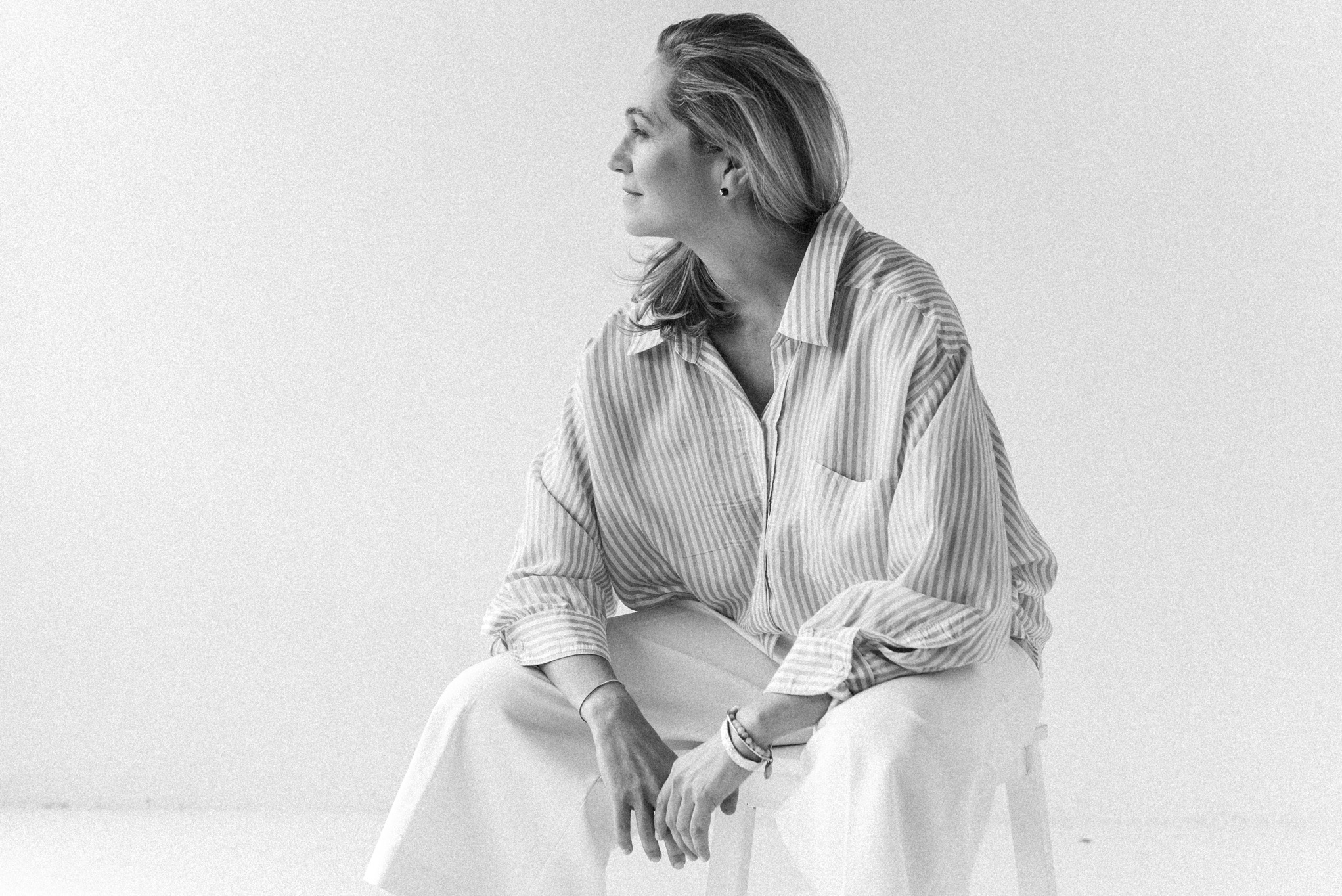 Czarno-białe zdjęcie kobiety w paski koszuli i białych spodniach, siedzącej bokiem na krześle na jasnym tle, z profilu widoczna twarz i zamyślone spojrzenie.