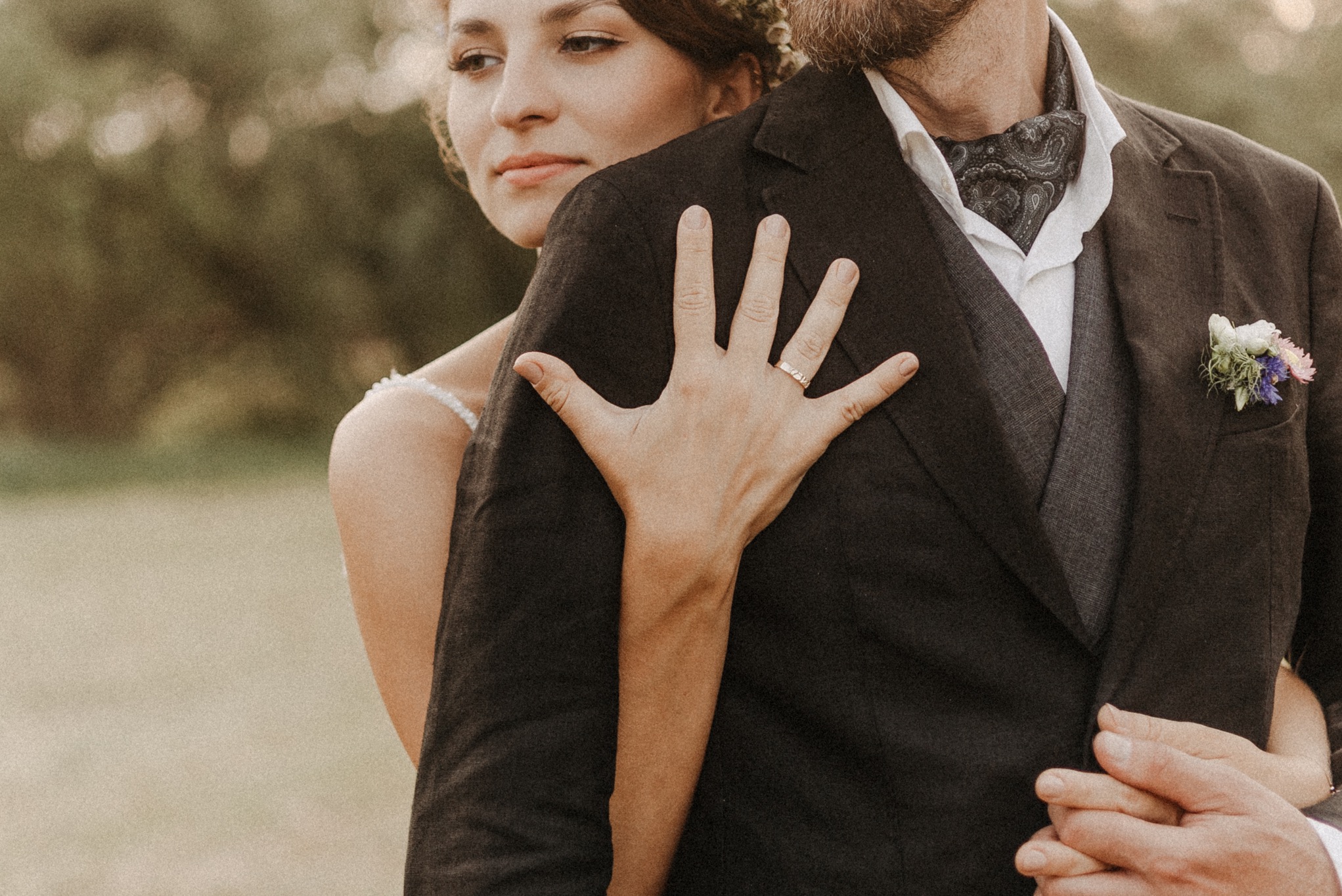 Panna młoda obejmuje pana młodego, dłoń z obrączką na ramieniu marynarki, w tle rozmyta zieleń.