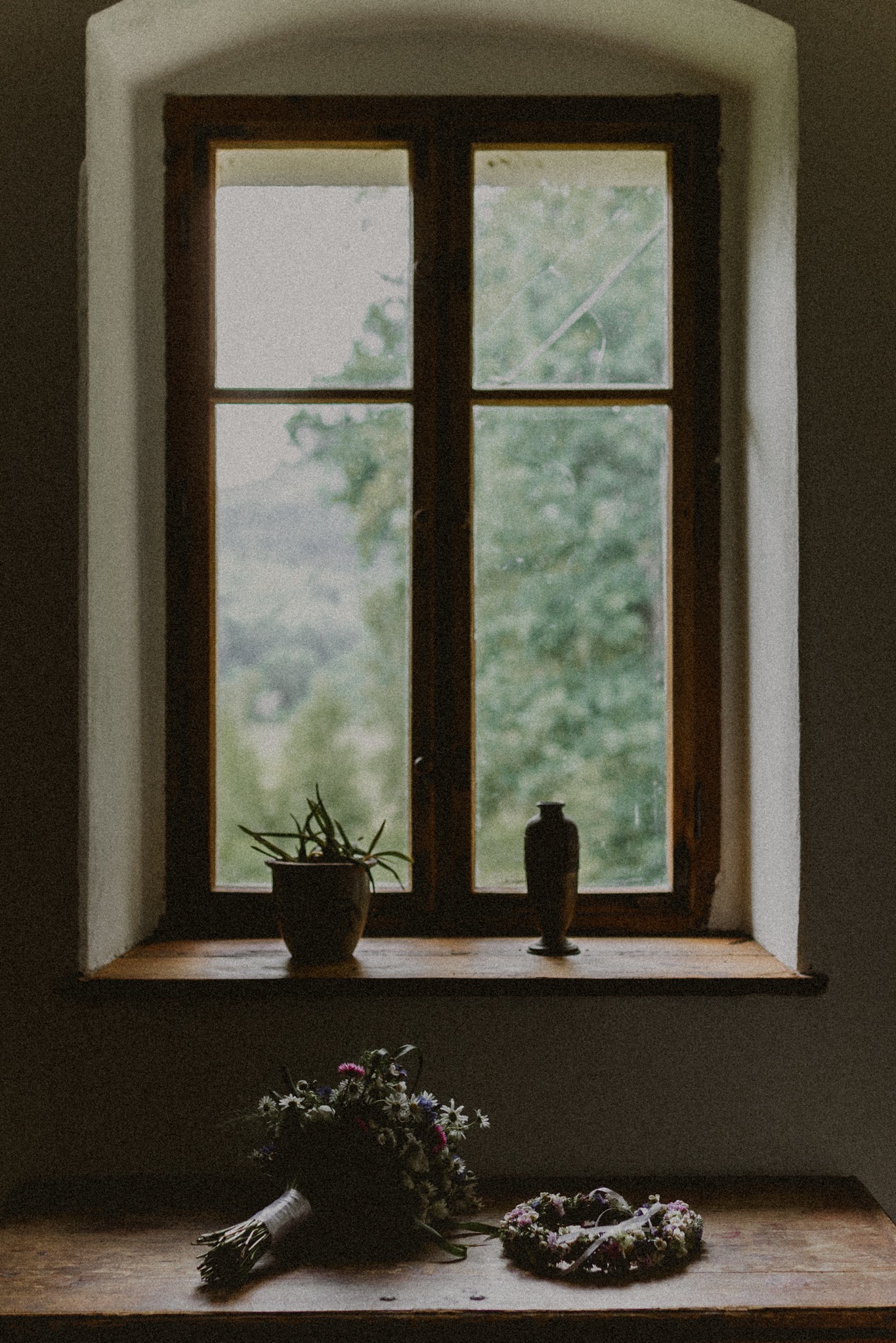 Bukiet ślubny i wianek leżące na drewnianym stole pod oknem w rustykalnym stylu, w tle widok na zieleń.
