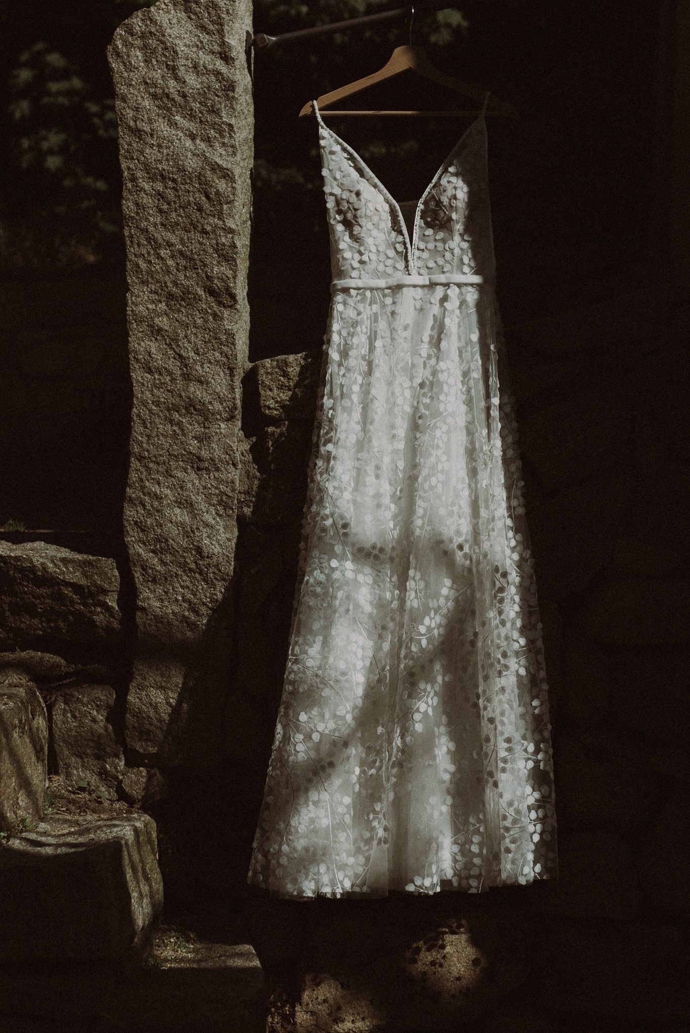 Delikatna suknia ślubna z motywem kwiatowym zawieszona na drewnianym wieszaku, na tle kamiennej ściany w cieniu, z subtelnym światłem padającym na materiał.