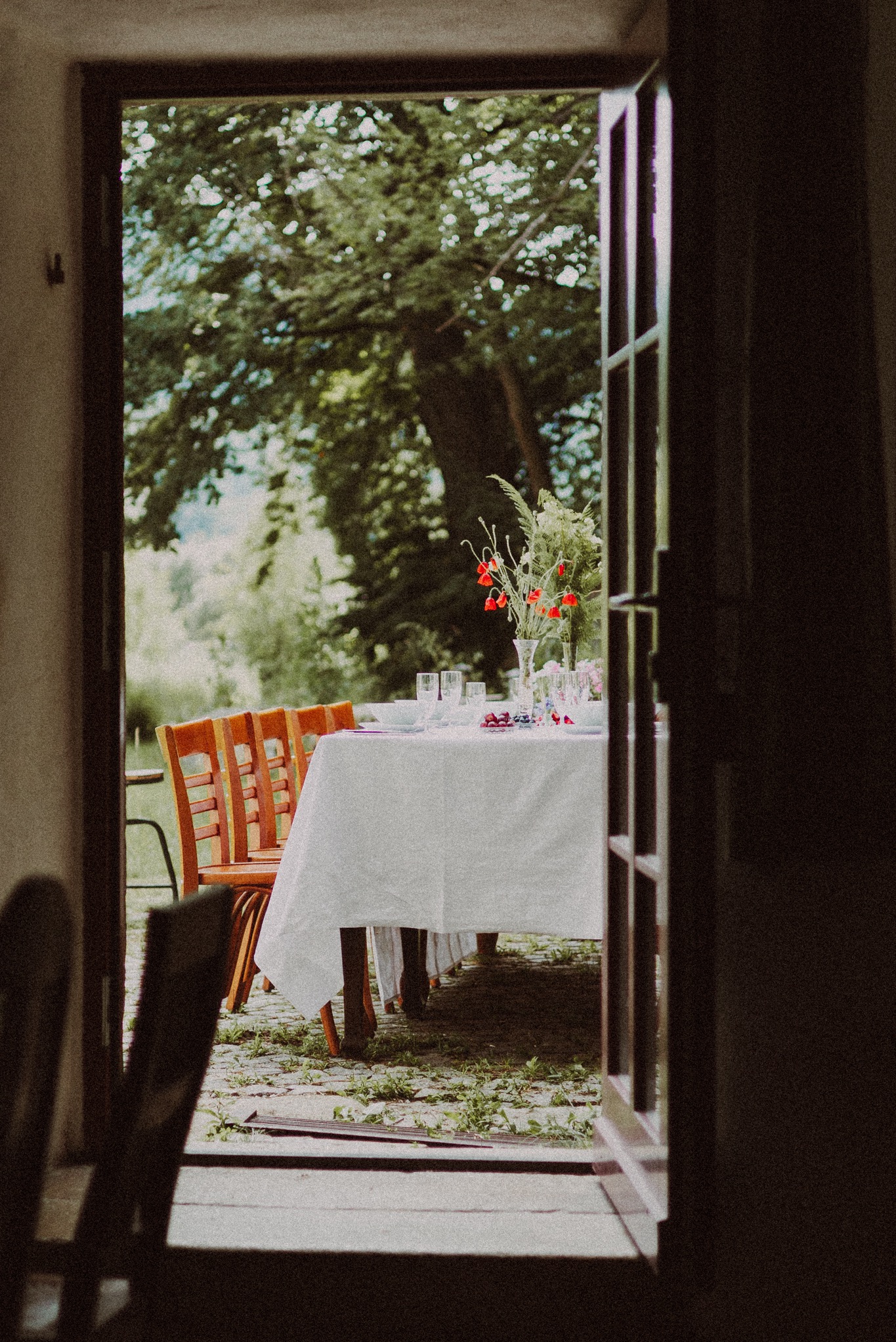 Ustawiony stół z białym obrusem, kwiatami w wazonie i nakryciami, widziany przez otwarte drzwi na zewnątrz, gdzie widać drzewa i trawnik.