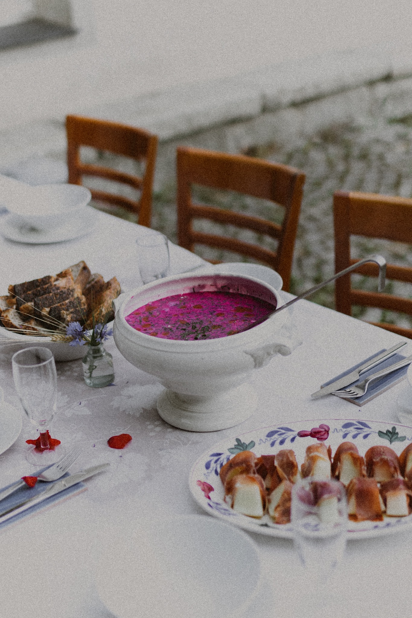 Uroczysty stół z chłodnikiem litewskim w wazie, chlebem, smalcem i dekoracjami kwiatowymi, przygotowany na zewnątrz.