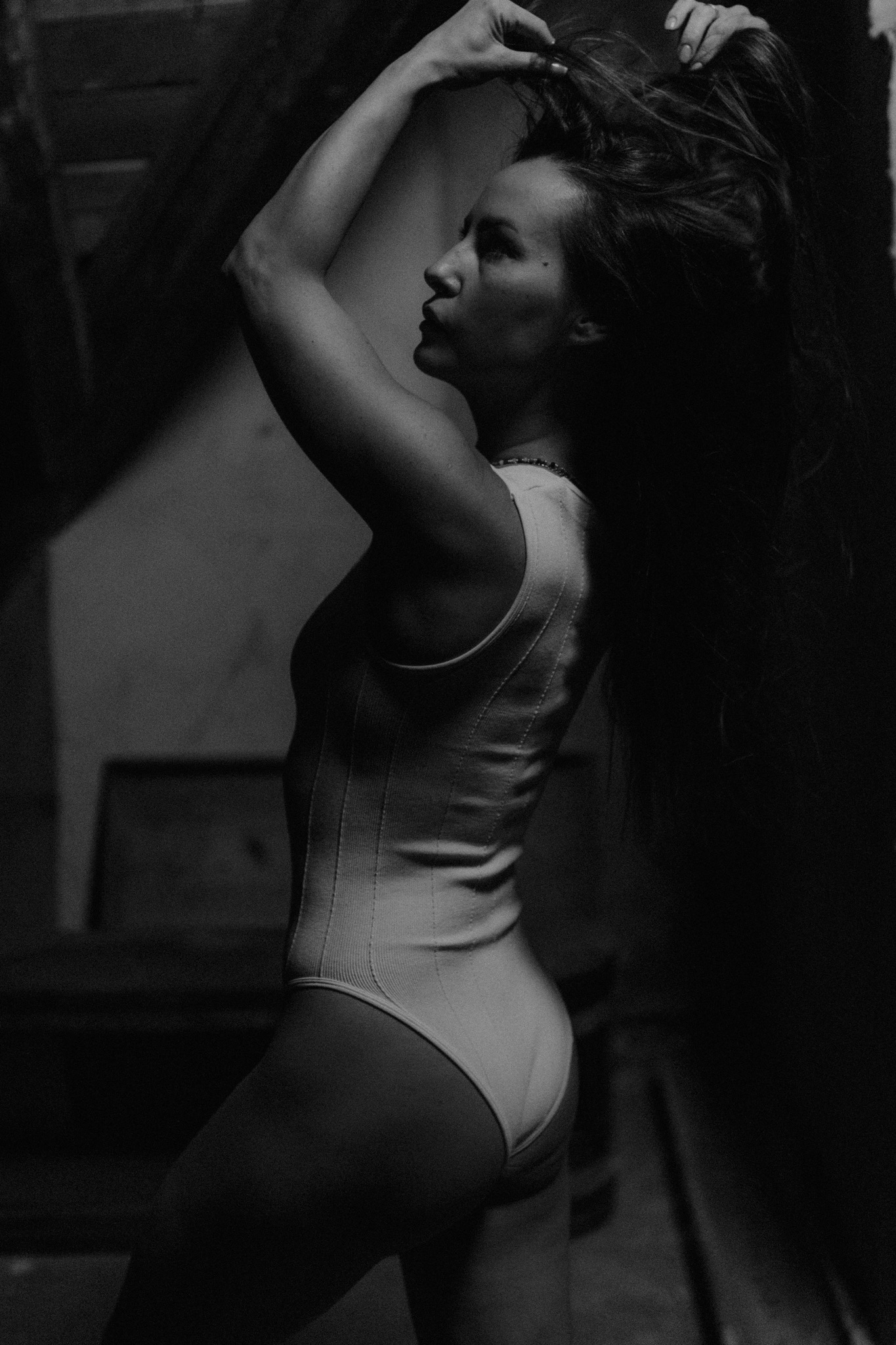 Czarno-białe zdjęcie kobiety w body, z rękami uniesionymi do góry, układającej włosy na tle schodów w ciemnym pomieszczeniu.