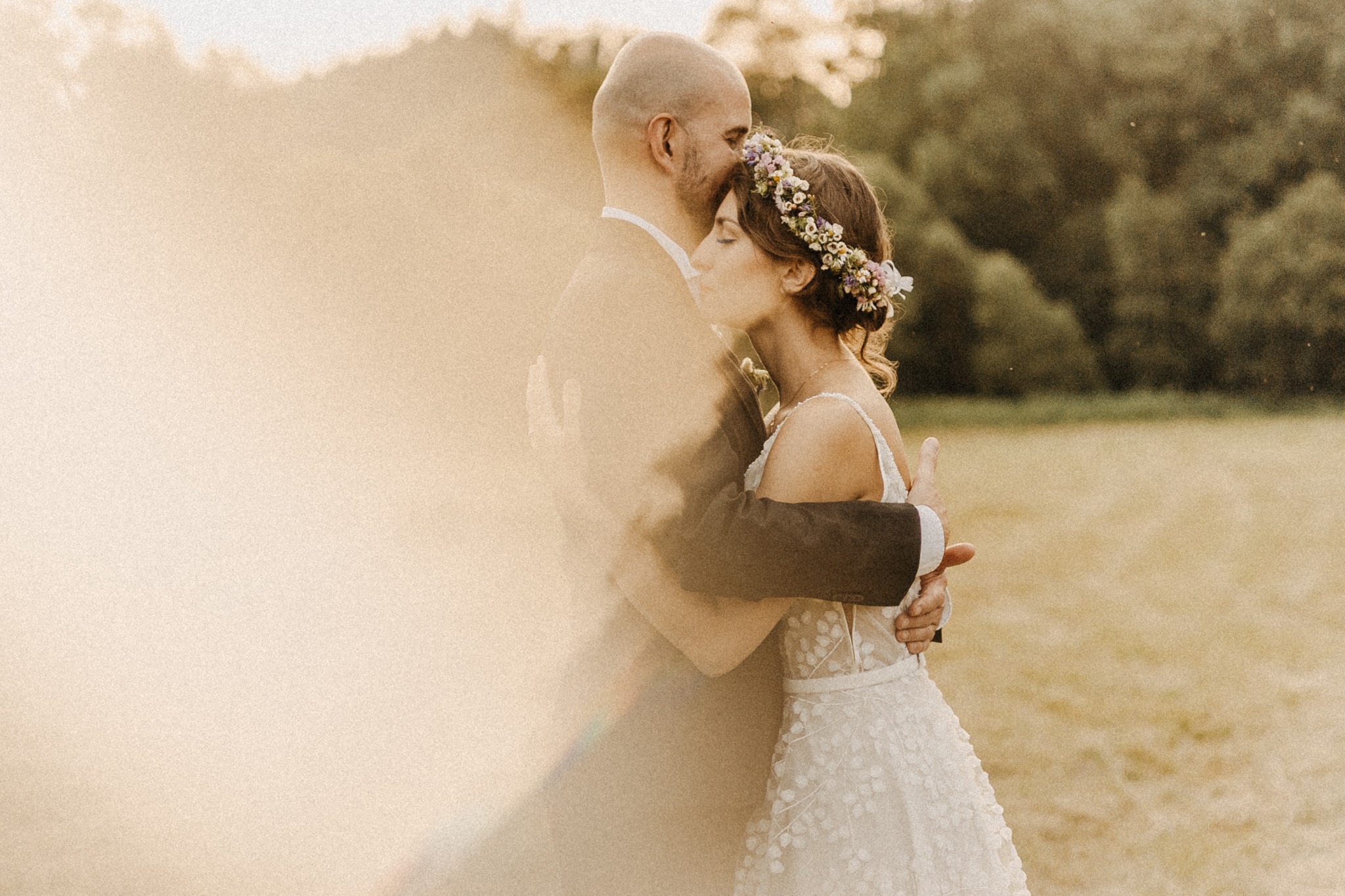 Para młoda w objęciach na tle zielonego pola, panna młoda w koronie z kwiatów, delikatne światło słoneczne