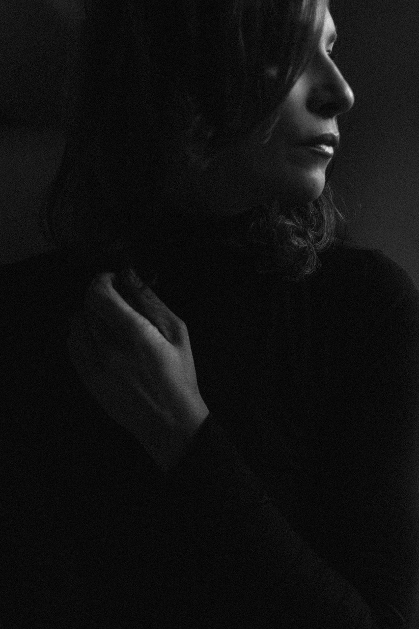 Czarno-białe portretowe ujęcie kobiety z delikatnym makijażem, dłoń dotyka szyi, skierowana w prawo, w ciemnym otoczeniu.