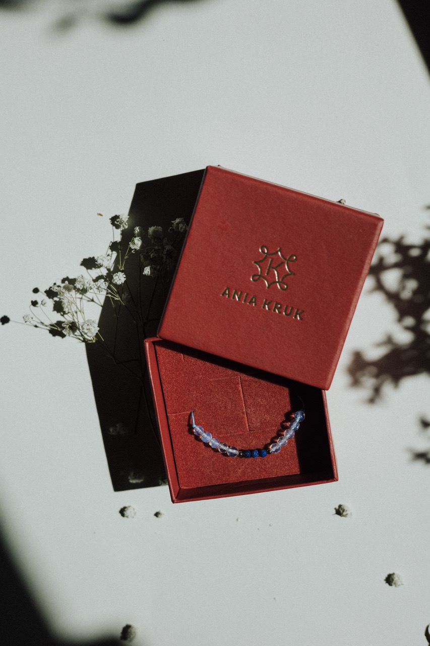 Bransoletka z niebieskich i opalizujących koralików w otwartym, czerwonym pudełku marki Ania Kruk, delikatne białe kwiaty i cienie na białym tle.