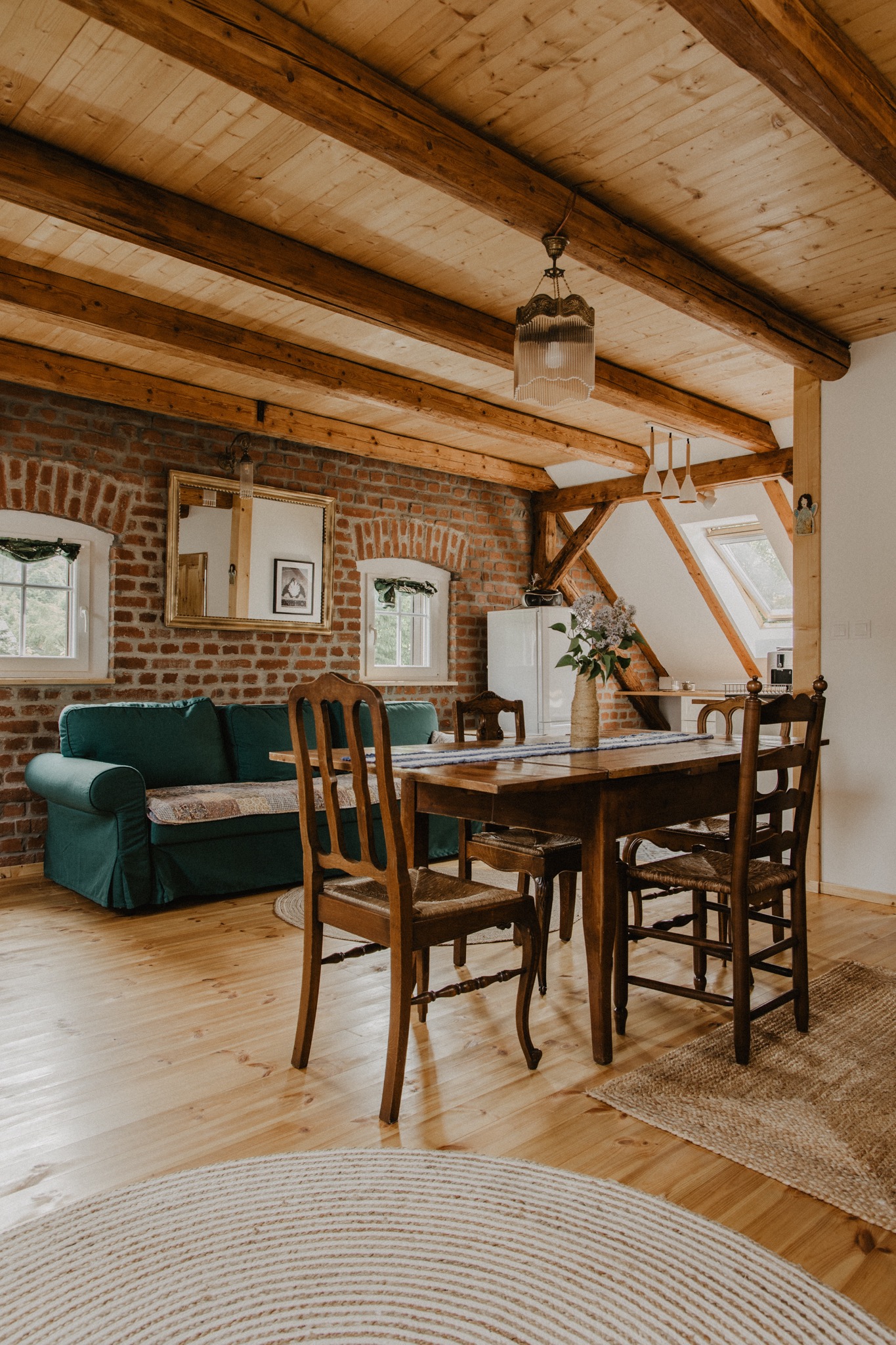 Rustykalne wnętrze: ceglana ściana, drewniany strop, zielona sofa, stół z bukietem bzu i okrągły jutowy dywan.