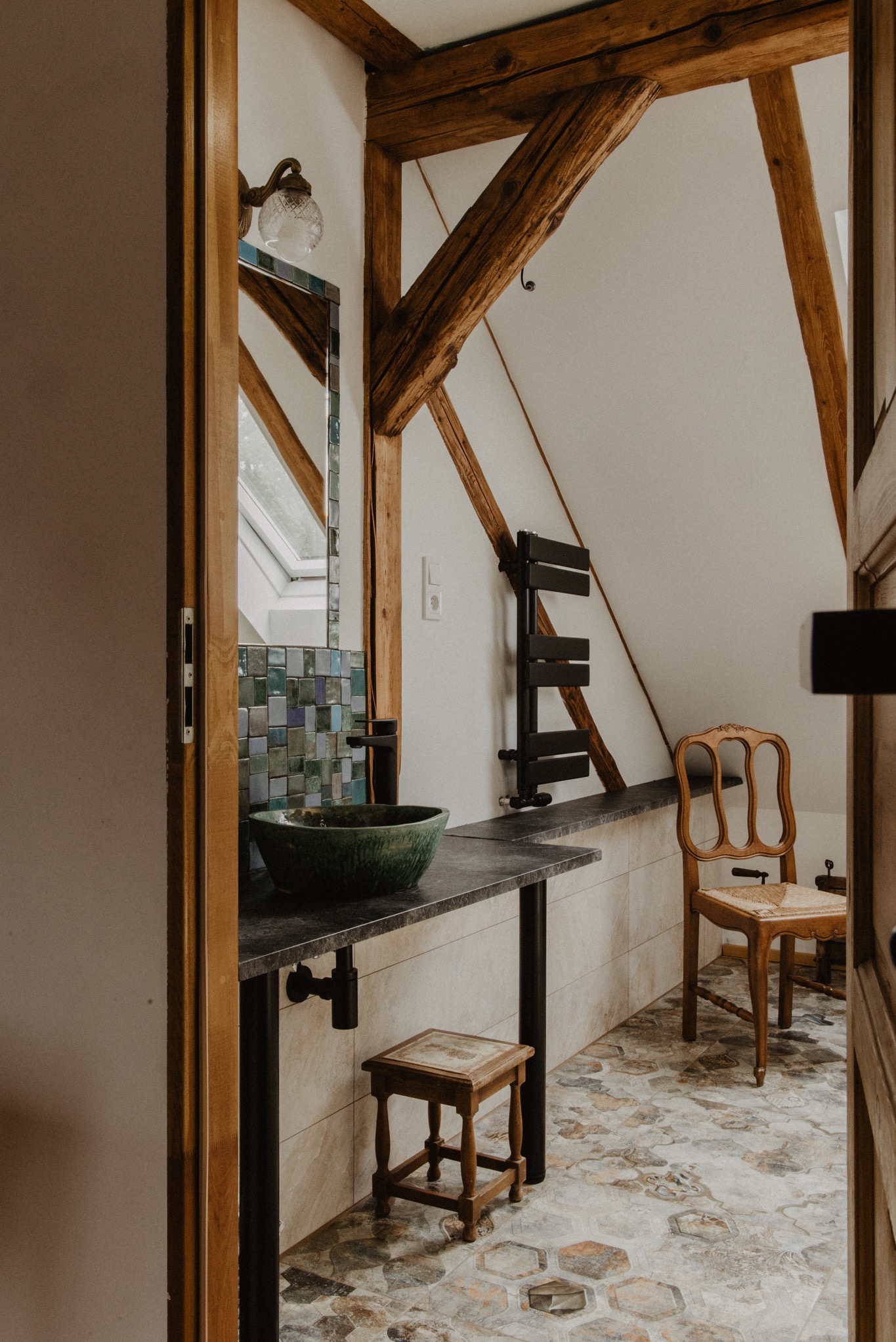 Rustykalna łazienka na poddaszu z drewnianymi belkami stropowymi, umywalką w odcieniach zieleni, lustrem w ramie z mozaiki i kamienną posadzką w kształcie plastra miodu.