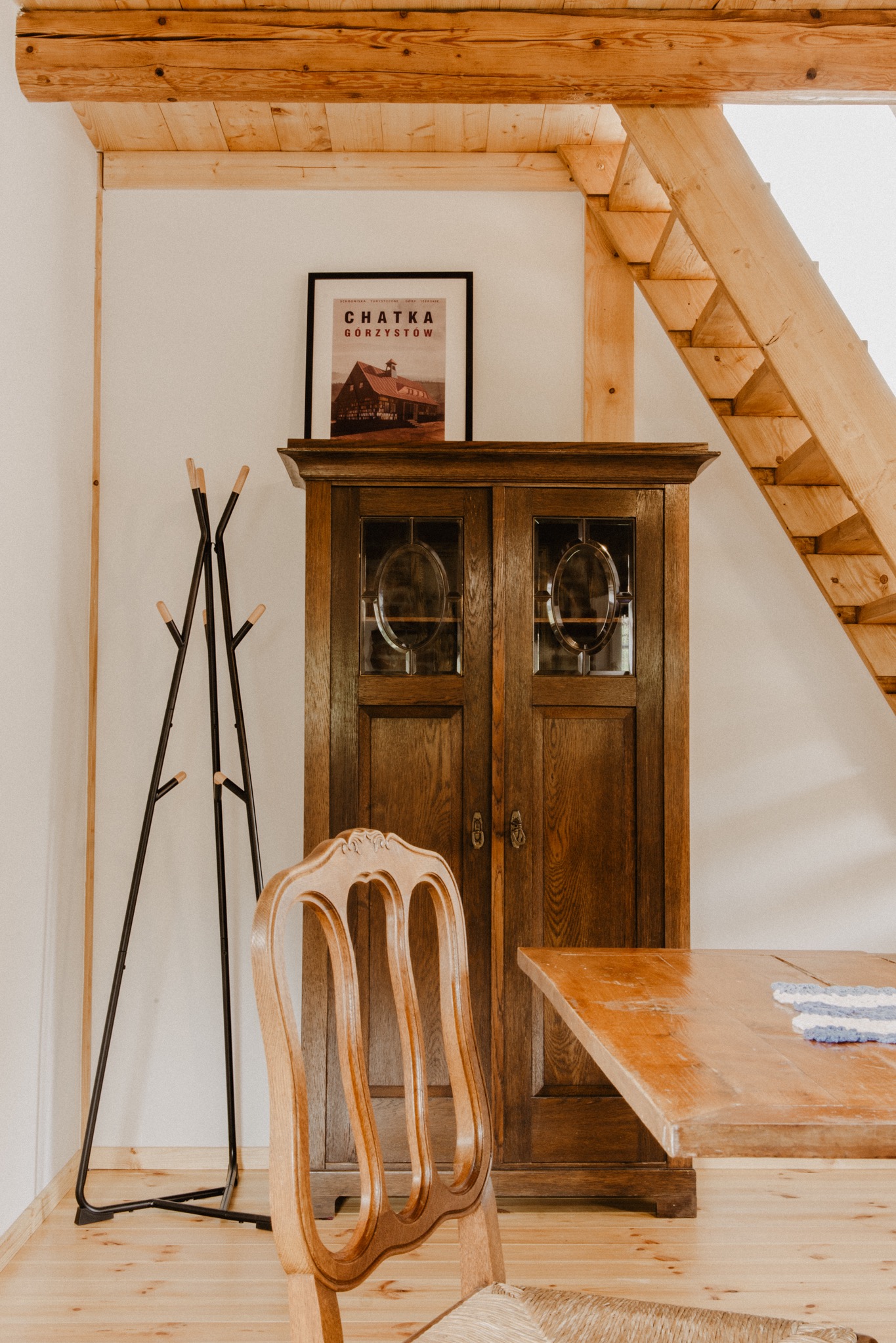 Rustykalne wnętrze: drewniana szafa z przeszkleniami, krzesło z plecionym siedziskiem, wieszak na ubrania i schody z belek, plakat 'Chatka Gorzystów' na szafie.