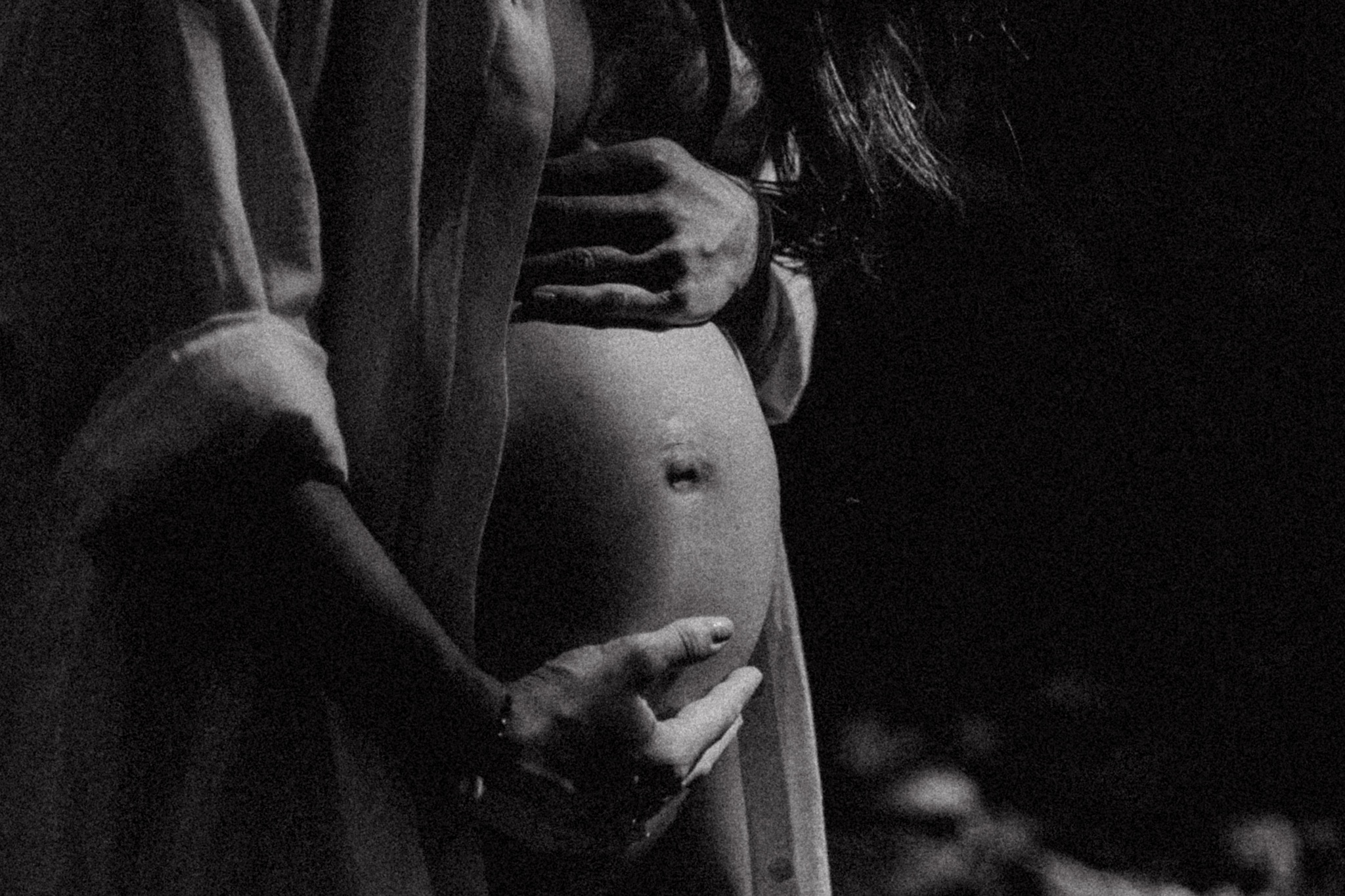Czarno-białe zdjęcie profilowe kobiety w ciąży, z rękami obejmującymi brzuch, w rozpiętej koszuli, z delikatnym oświetleniem podkreślającym kształt brzucha.
