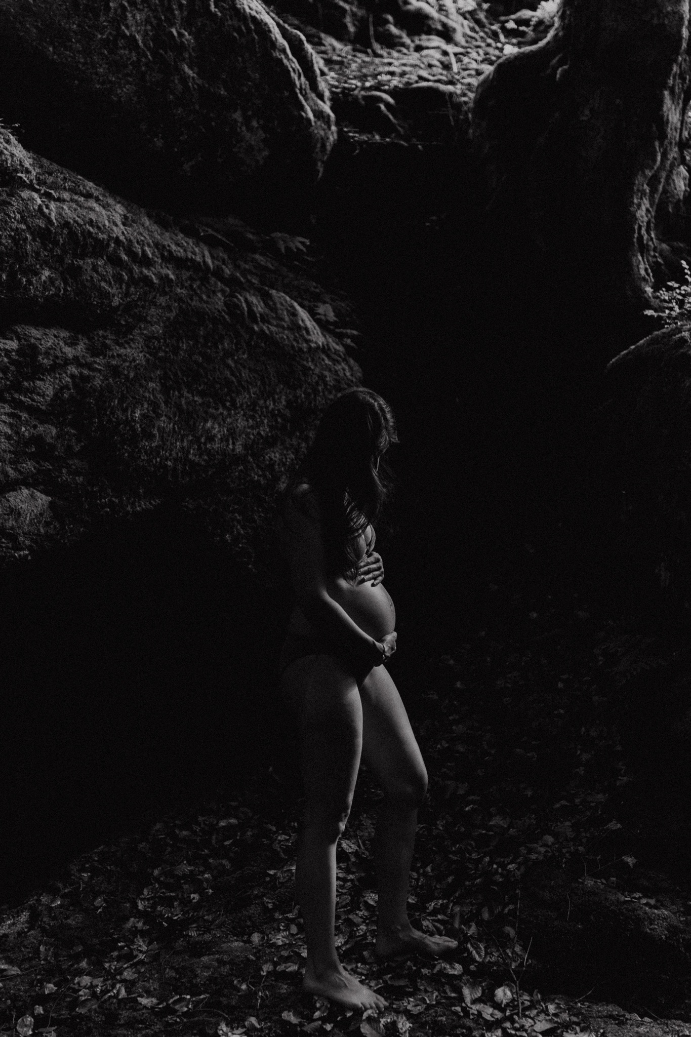 Czarno-białe zdjęcie kobiety w ciąży, stojącej boso w otoczeniu skał i liści, z rękami na brzuchu, w naturalnym oświetleniu.