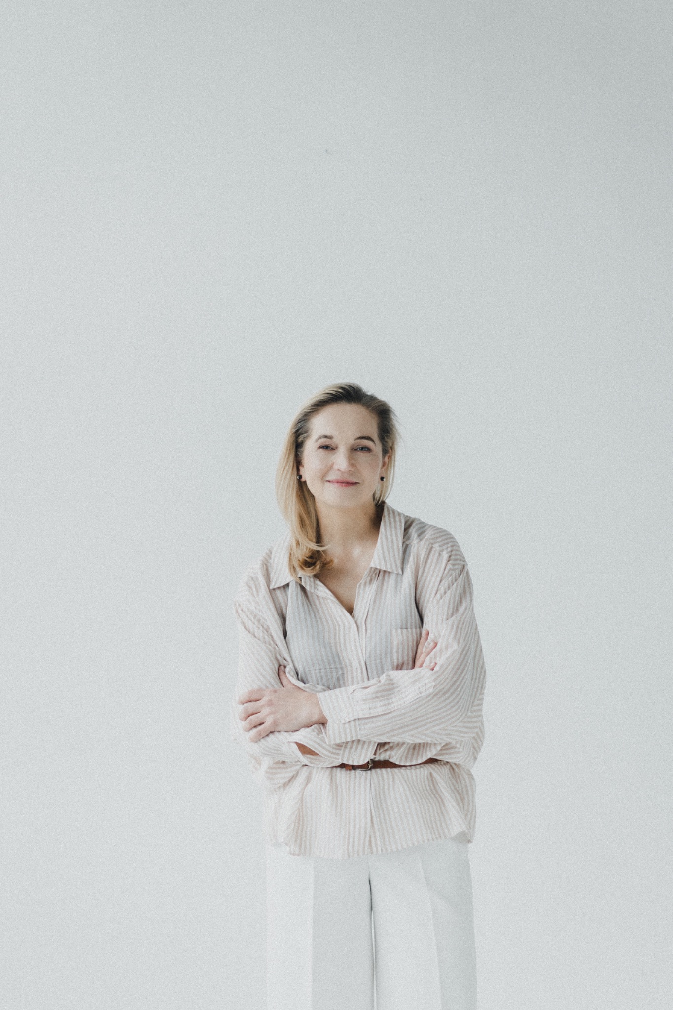 Portret kobiety z blond włosami w koszuli w paski i białych spodniach, stojącej na jasnym tle z założonymi rękami.