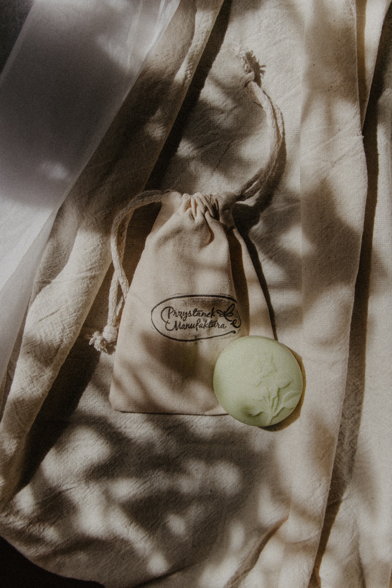 Naturalne mydło w pastelowym odcieniu zieleni leży obok lnianego woreczka z logo 'Przystanek Manufaktura' na tle beżowej tkaniny w cieniu.