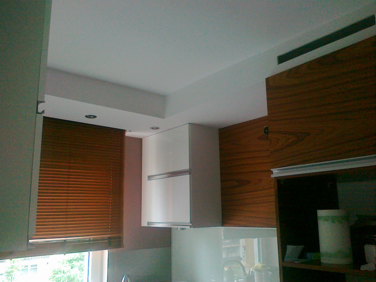 Fragment wykończonej kuchni z drewnianymi i białymi szafkami wiszącymi, brązowymi żaluzjami okiennymi i oświetleniem punktowym w suficie podwieszanym.