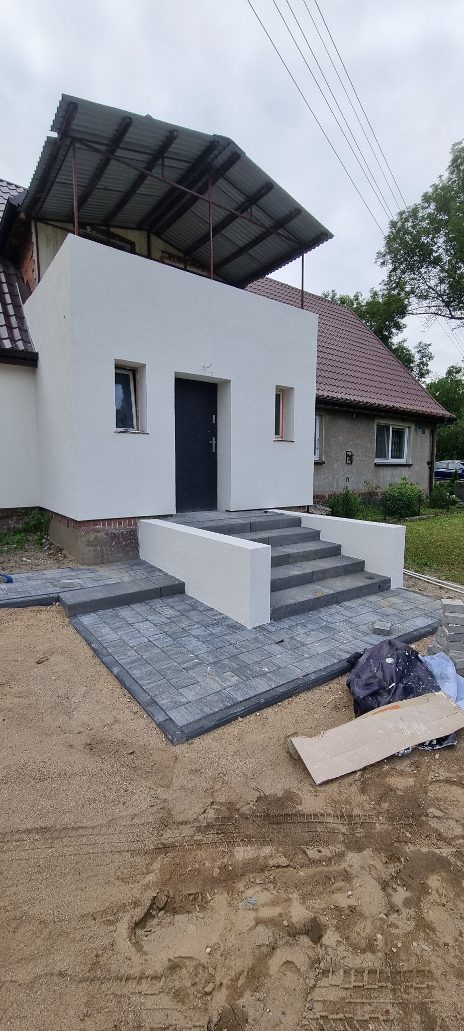 Nowo wyłożona szara kostka brukowa prowadząca do wejścia do budynku z białym otynkowanym gankiem i schodami, widoczne ślady opon na piasku.