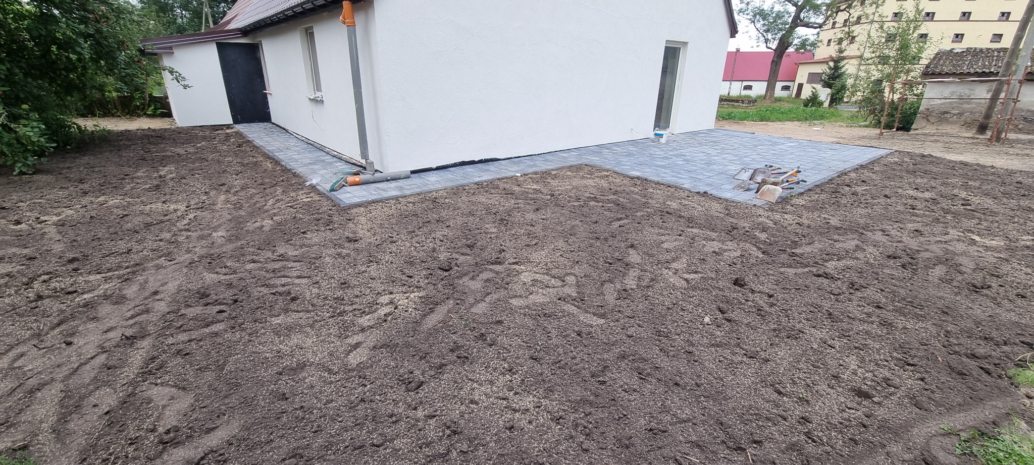 Szara kostka brukowa ułożona przy białym budynku z czarnymi drzwiami, widoczne narzędzia i przygotowany teren pod trawnik.