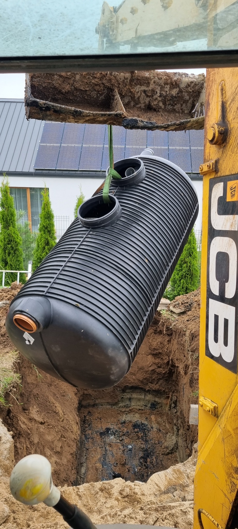Czarny, żebrowany zbiornik na deszczówkę zawieszony na pasach nad wykopem przy użyciu żółtej koparki JCB, w tle dom z panelami słonecznymi i zielone tuje.