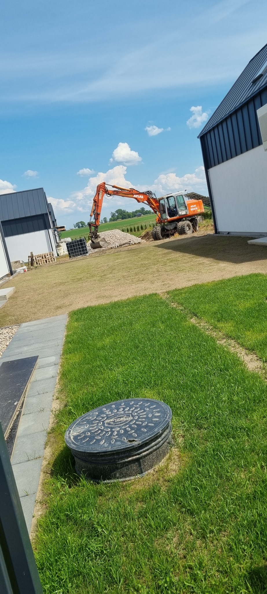 Pomarańczowa koparka Atlas 1504M pracuje na terenie budowy, przygotowując teren pod trawnik i układanie kostki, widoczny właz kanalizacyjny.