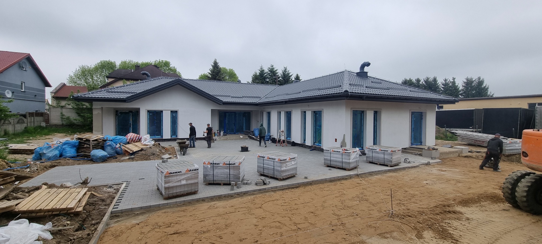 Nowoczesny dom jednorodzinny w trakcie budowy, z ułożoną częścią podjazdu z szarej kostki brukowej, palety z kostką Durabet czekają na ułożenie, robotnicy pracują na zewnątrz, a w tle widoczny...