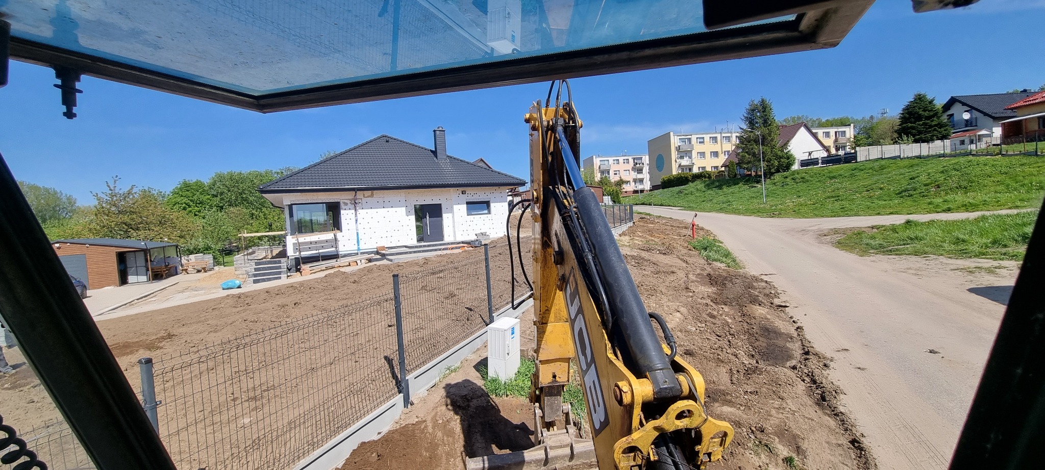Widok z kabiny koparki na nowo postawiony dom w trakcie ocieplania i montażu ogrodzenia panelowego z podmurówką, przygotowany teren pod trawnik.
