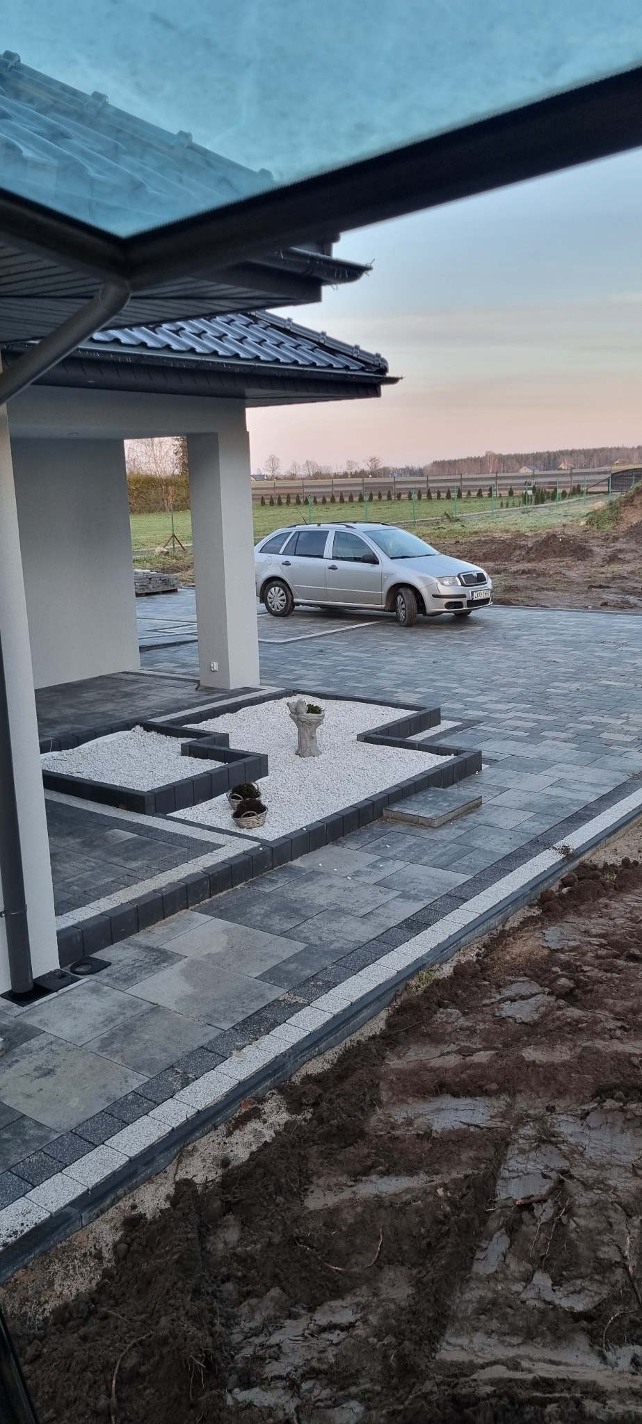 Nowo ułożona szara kostka brukowa z białym grysem tworząca dekoracyjne rabaty przed domem z zaparkowanym srebrnym samochodem.