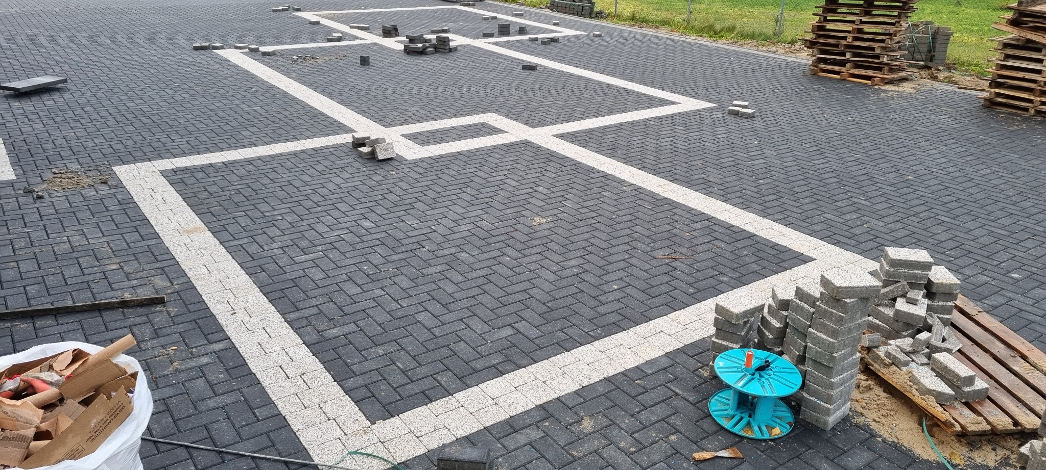 Częściowo ułożona, czarno-biała kostka brukowa w jodełkę, wyznaczająca kształt podjazdu, z paletami kostki i niebieską szpulą kabla w tle.
