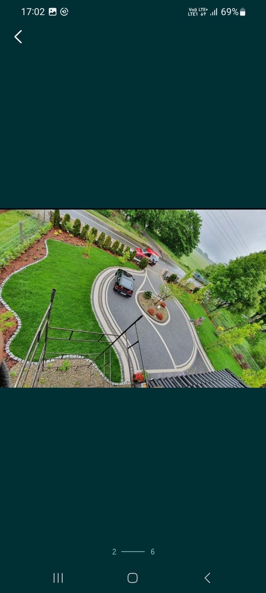 Widok z góry na podjazd z kostki brukowej w kształcie owalu z centralną wyspą zieleni i ozdobnymi krzewami, otoczony trawnikiem z obrzeżem z kostki granitowej, w tle zaparkowane dwa samochody i droga.