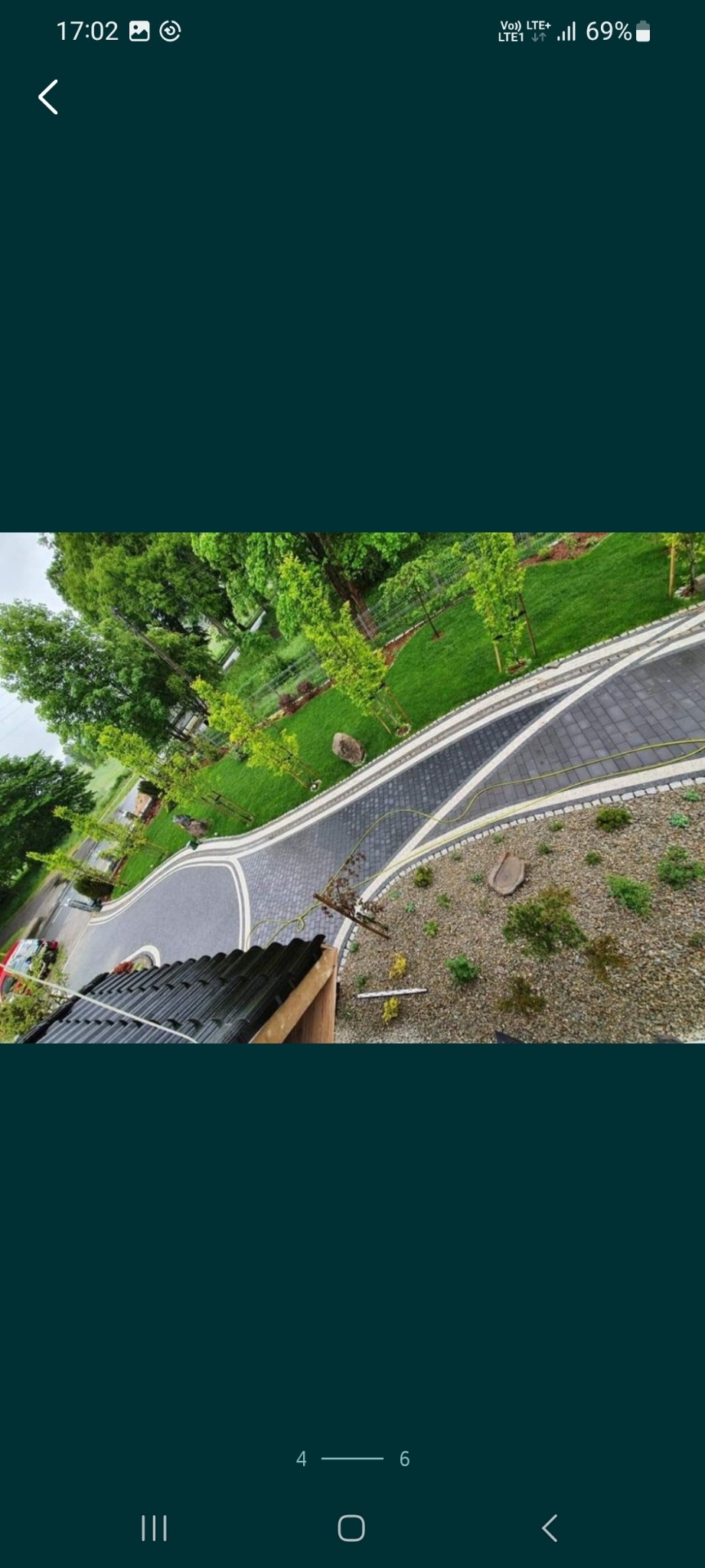 Czarno-szara kostka brukowa ułożona w łukowaty wzór otoczona zielenią trawnika i drzew, widok z góry z fragmentem dachu w kadrze.