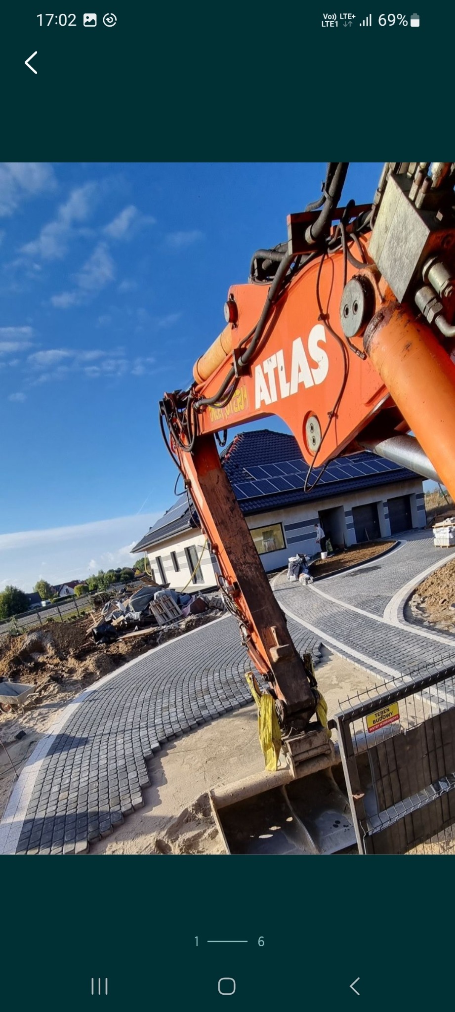 Pomarańczowa koparka Atlas na tle nieba i domu z panelami słonecznymi, widoczny fragment nowo ułożonej, łukowatej ścieżki z szarej kostki brukowej, teren budowy z tablicą ostrzegawczą.