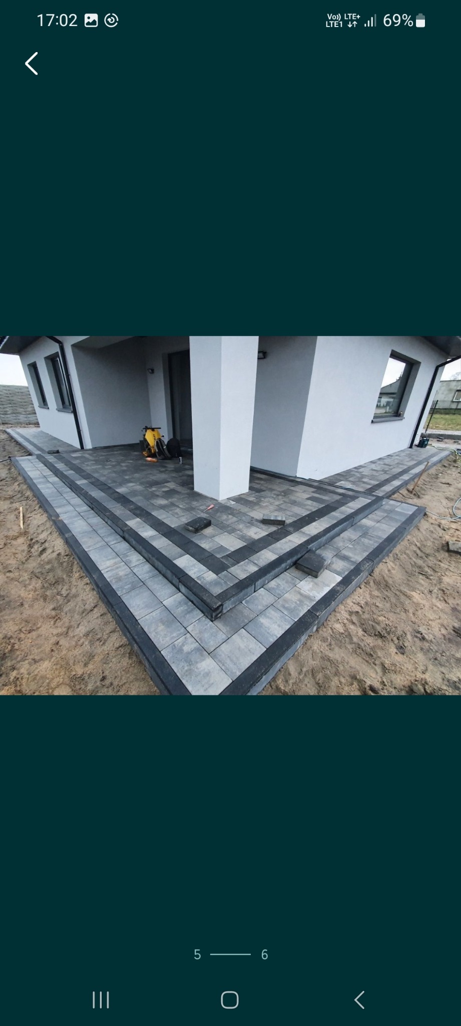 Ułożona kostka brukowa przed nowoczesnym domem z szarymi ścianami i białymi kolumnami, widoczne schody z kostki i częściowo ułożony chodnik na piaszczystym podłożu, w tle zagęszczarka.