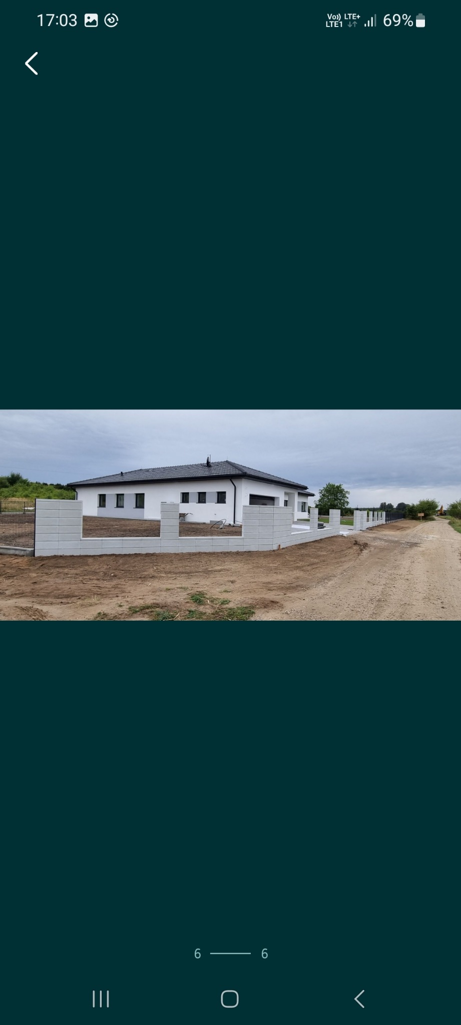 Nowoczesny dom jednorodzinny z białymi ścianami i ciemnym dachem, otoczony ogrodzeniem z betonowych prefabrykatów, widoczne przygotowanie terenu pod trawnik, droga gruntowa z boku działki.