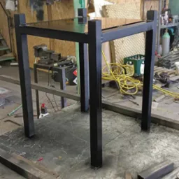 Czarny, metalowy stół o kwadratowym blacie i nogach, wykonany w warsztacie ślusarskim, widoczne narzędzia i materiały na stole roboczym.