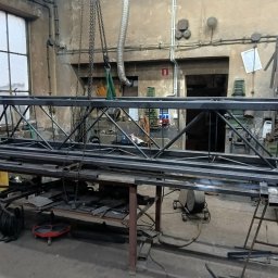 DON SERWIS Paweł Wójcik - Metalowa konstrukcja kratowa, prawdopodobnie w trakcie spawania, zawieszona nad stołem w warsztacie. Widoczne narzędzia i elementy metalowe w tle.