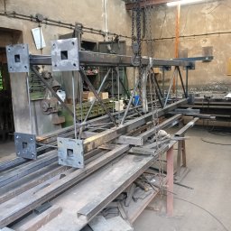 DON SERWIS Paweł Wójcik - Konstrukcja stalowa z kwadratowymi elementami, podwieszona na łańcuchu w warsztacie. Widoczne profile i elementy spawane. Surowe wykończenie, warsztatowe otoczenie.