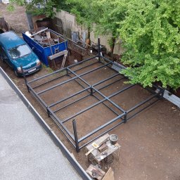 DON SERWIS Paweł Wójcik - Metalowa konstrukcja szkieletowa w ogrodzie, widok z góry. Na ziemi leżą narzędzia. W tle zaparkowany samochód i kontener ze złomem.