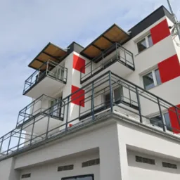 Nowoczesny budynek z balkonami z metalowymi balustradami w geometryczne wzory czerwieni i bieli. Widok z dołu w słoneczny dzień, podkreślający minimalistyczny design.