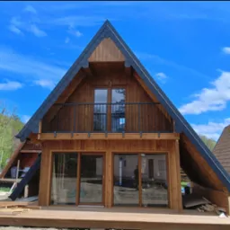 Nowoczesny, drewniany dom w stylu góralskim z balkonem i czarną balustradą na tle błękitnego nieba. Widoczne detale wykończenia dachu i drewniana elewacja.