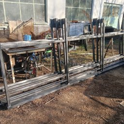 DON SERWIS Paweł Wójcik - Stalowe ramy konstrukcyjne z profili kwadratowych, ułożone warstwami na ziemi, w tle warsztat z narzędziami i maszynami, prawdopodobnie gotowe elementy do montażu ogrodzenia.