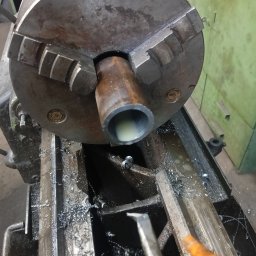 DON SERWIS Paweł Wójcik - Metalowa rura w uchwycie tokarskim podczas obróbki, widoczne wióry i narzędzie skrawające. Szare kolory maszyny, chłodziwo wewnątrz rury, zbliżenie na proces.