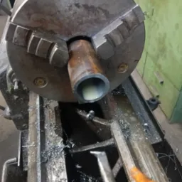 Metalowa rura w uchwycie tokarskim podczas obróbki, widoczne wióry i narzędzie skrawające. Szare kolory maszyny, chłodziwo wewnątrz rury, zbliżenie na proces.