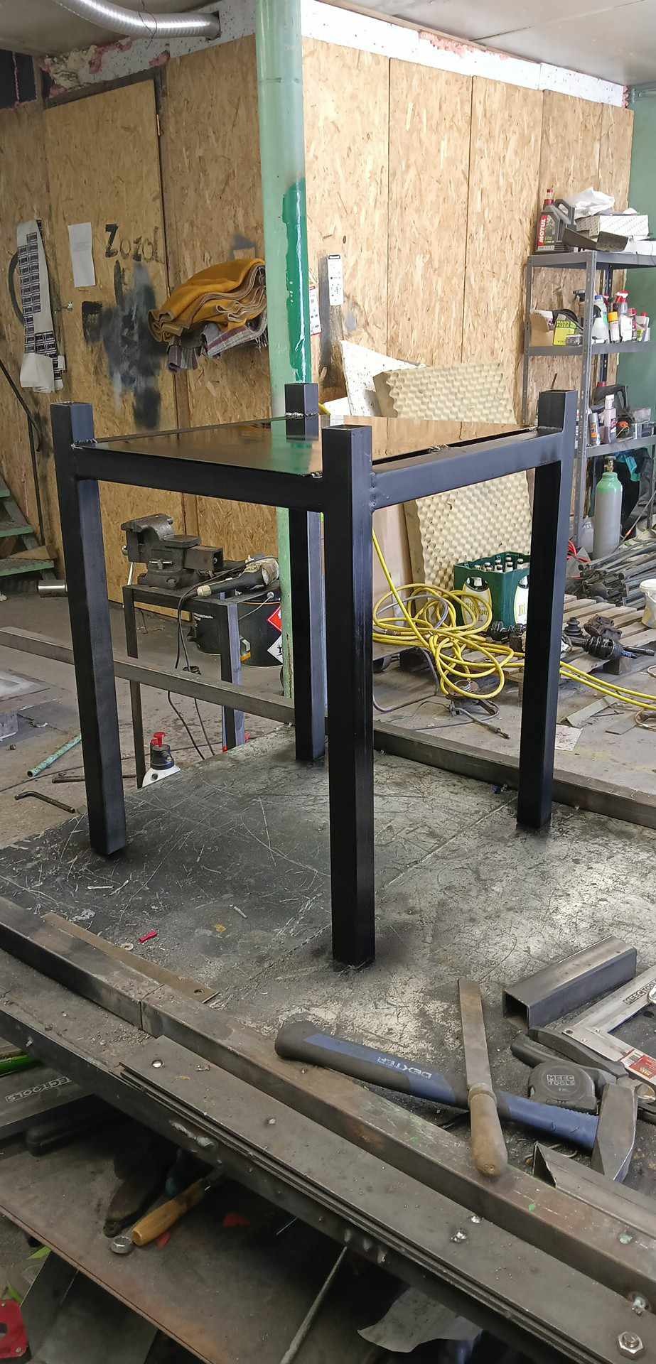 Czarny, metalowy stół o kwadratowym blacie i nogach, wykonany w warsztacie ślusarskim, widoczne narzędzia i materiały na stole roboczym.