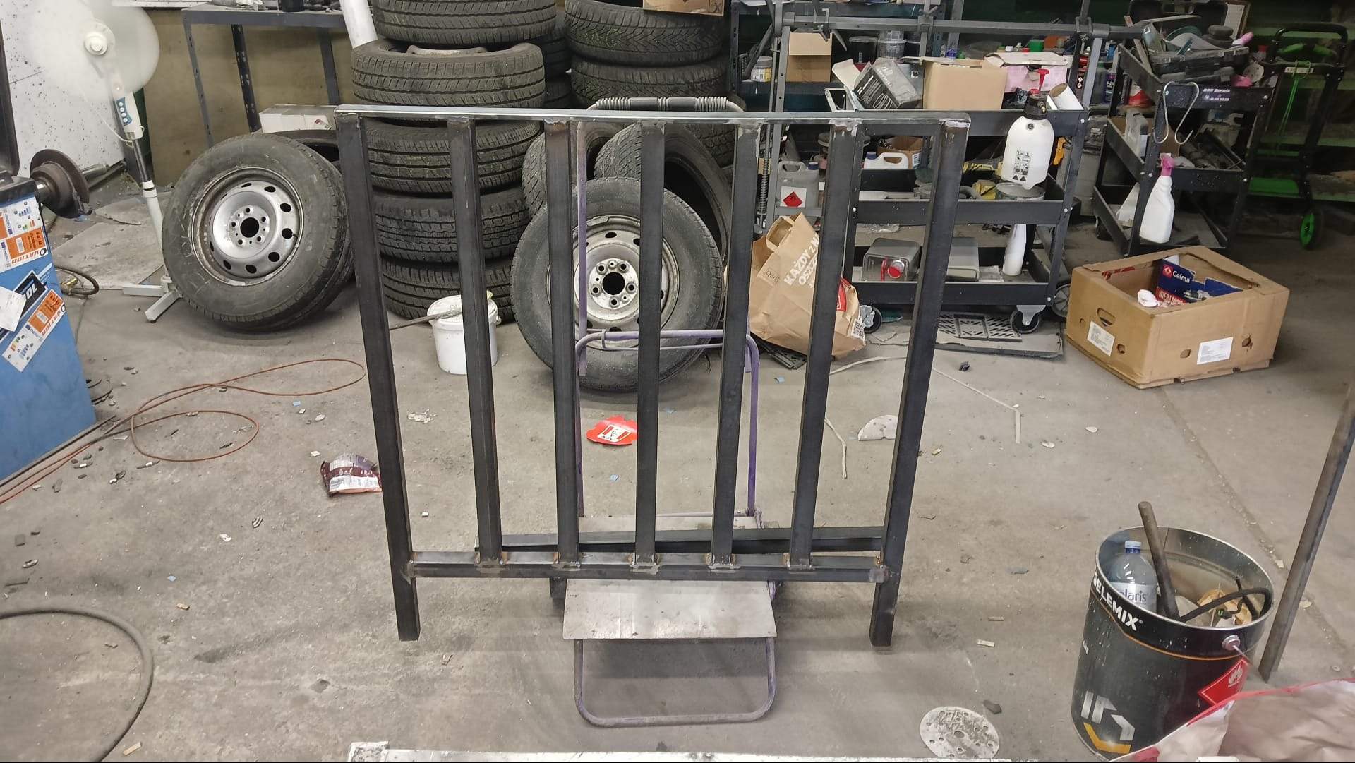 Surowa, spawana konstrukcja metalowa, przypominająca fragment balustrady lub ogrodzenia, na tle opon samochodowych i warsztatowego nieładu.